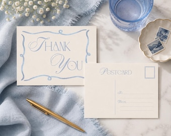 Pode incluir: Um cartão de agradecimento cor de creme com uma borda em aguarela azul-claro e o texto Thank You, ao lado de um postal a condizer com a palavra Postcard, uma caixa para selo e linhas para endereço, com uma caneta dourada sobre tecido azul.