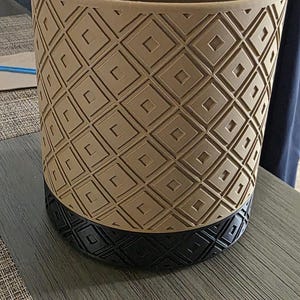 May include: Tan and black decorative planter with a geometric diamond pattern. The planter has a cylindrical shape and is made of a durable material. The base is black, and the upper portion is tan.