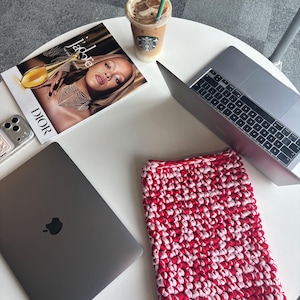 May include: A white table displays a laptop, a magazine, a phone, and an iced coffee. A red and white crocheted laptop sleeve is also on the table. The laptop is closed, showing the Apple logo.