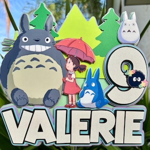 May include: A colorful cake topper featuring characters from the animated film, My Neighbor Totoro. The design includes a large gray Totoro, a girl with a red umbrella, a blue Totoro, and the name "VALERIE" with the number "9".