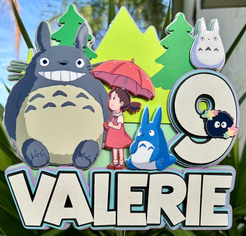 May include: A colorful cake topper featuring characters from the animated film, My Neighbor Totoro. The design includes a large gray Totoro, a girl with a red umbrella, a blue Totoro, and the name "VALERIE" with the number "9".