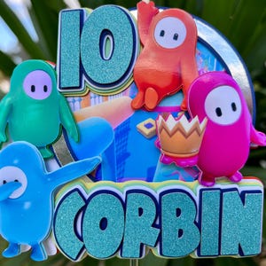 May include: A layered cake topper featuring the number 10 and the name CORBIN in glittery teal letters. The design includes four colorful cartoon characters in green, orange, blue, and pink, with one character holding a brown crown.