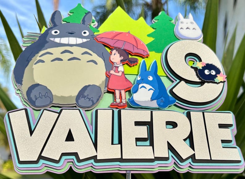 May include: A colorful cake topper featuring characters from the animated film "My Neighbor Totoro." The design includes the characters Totoro, Mei, and the Catbus, along with the name "VALERIE" in large, layered letters. The number "9" is also included.