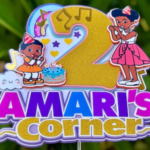 以下が含まれることがあります： 紫と金色の「AMARI'S CORNER」の文字が入った、カラフルなバースデーケーキトッパー。数字の「2」が大きく表示され、女の子、音符、虹、バースデーケーキの漫画イラストが添えられています。