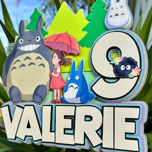May include: A layered cake topper featuring characters from the animated film, "My Neighbor Totoro." The design includes a large gray Totoro, a girl with an umbrella, and the number "9" with the name "Valerie". The background has green trees and a light green mountain.