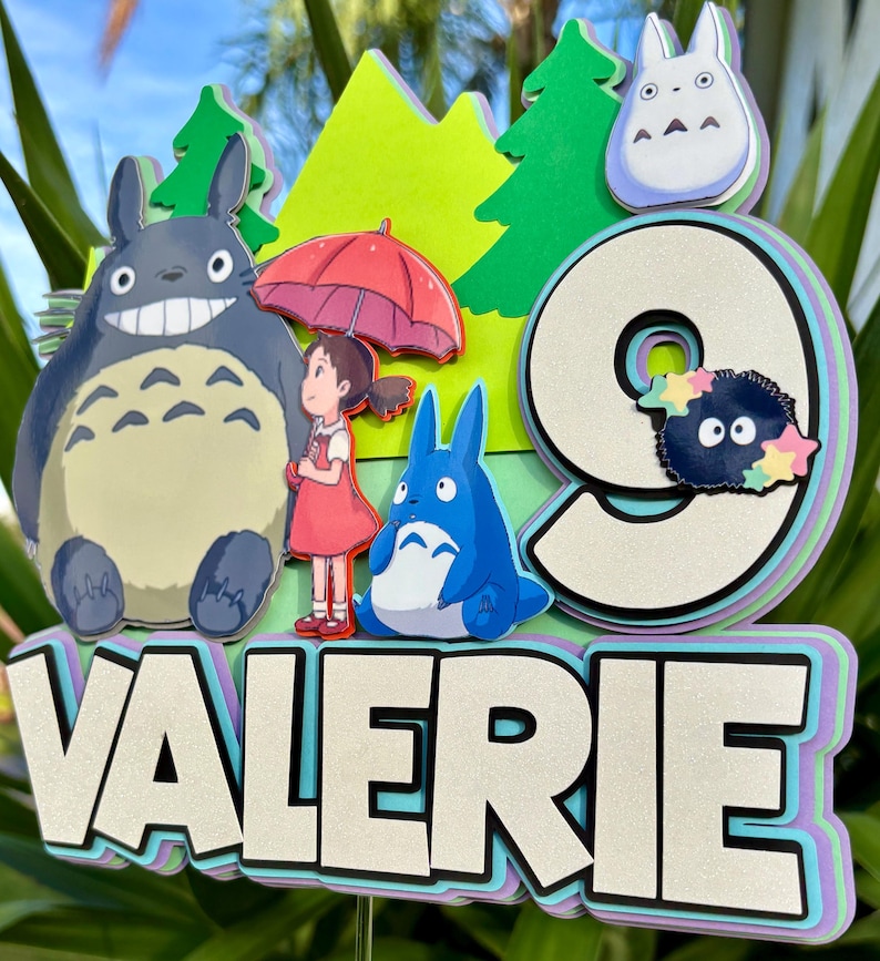 May include: A layered cake topper featuring characters from the animated film, "My Neighbor Totoro." The design includes a large gray Totoro, a girl with an umbrella, and the number "9" with the name "Valerie". The background has green trees and a light green mountain.