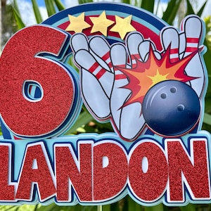 May include: A colorful bowling-themed birthday decoration featuring the number "6" in red glitter, bowling pins, a bowling ball, and the name "LANDON" in red glitter. The design includes gold stars and a blue, red, and white color scheme.