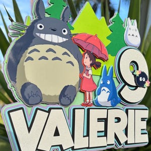 May include: A layered cake topper featuring characters from the animated film, My Neighbor Totoro. The design includes Totoro, a girl with an umbrella, and the name "VALERIE" with a number "9". The color palette includes shades of gray, blue, green, and red.