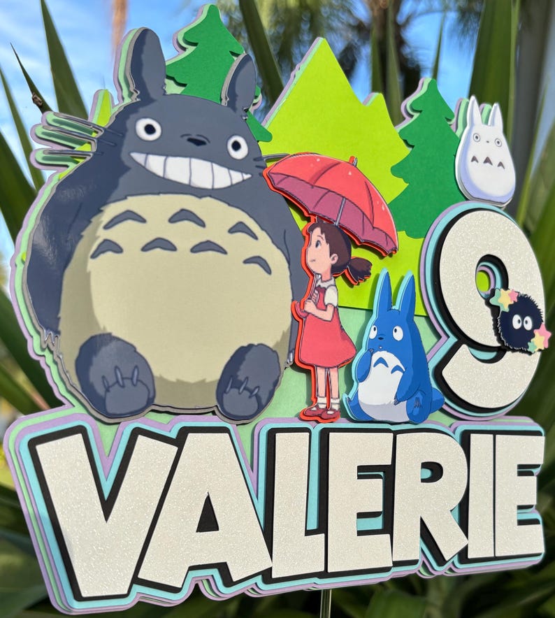 May include: A layered cake topper featuring characters from the animated film, My Neighbor Totoro. The design includes Totoro, a girl with an umbrella, and the name "VALERIE" with a number "9". The color palette includes shades of gray, blue, green, and red.