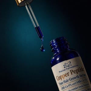 May include: A blue glass bottle of Precise Formulations Copper Peptide True Hair Growth Serum with a gold-capped dropper. A single drop of the dark blue liquid is suspended from the glass pipette, positioned above the open bottle neck.