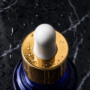 May include: A dark blue glass bottle featuring a metallic gold collar and a white rubber dropper bulb. The top of the bottle is covered in small water droplets, reflecting light across the gold surface and the smooth, rounded white dropper tip.