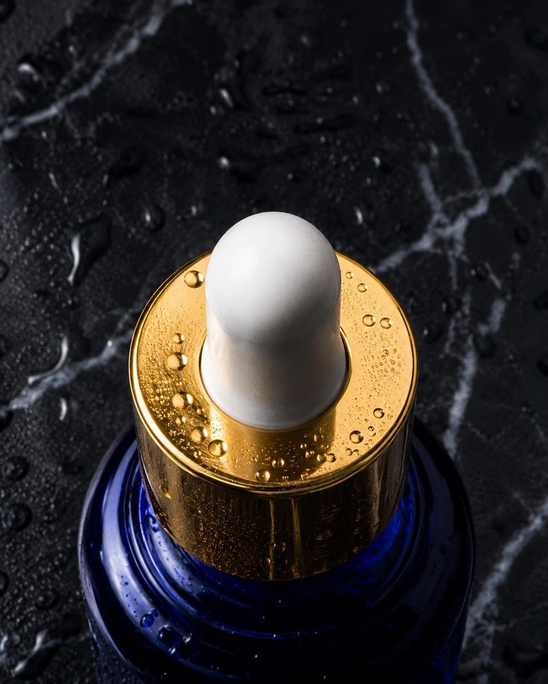 May include: A dark blue glass bottle featuring a metallic gold collar and a white rubber dropper bulb. The top of the bottle is covered in small water droplets, reflecting light across the gold surface and the smooth, rounded white dropper tip.