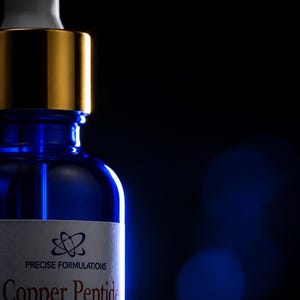 May include: A blue glass dropper bottle with a gold collar and white dropper top. The label reads PRECISE FORMULATIONS Copper Peptide True Hair Growth Serum GHK-Cu 6% and AHK-Cu 1% (with Hyaluronic Acid) with a circular atom-like logo above.