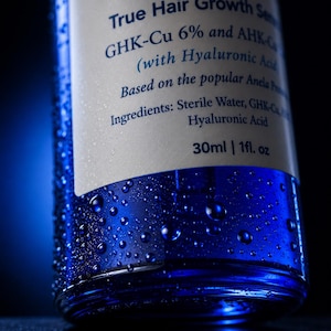 May include: A blue glass bottle of Copper Peptide True Hair Growth Serum with a white label. The label lists GHK-Cu 6% and AHK-Cu, Hyaluronic Acid, and ingredients including Sterile Water. The bottle contains 30ml or 1 fl. oz and is covered in water droplets.