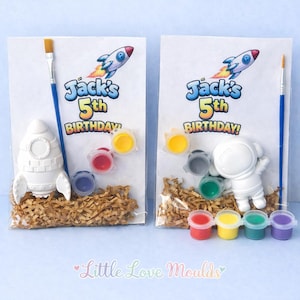 Puede incluir: Dos kits de manualidades para pintar para una fiesta de cumpleaños. Cada kit incluye un cohete o astronauta de cerámica blanca, botes de pintura, un pincel y una bolsa de papel triturado. Las bolsas tienen el texto "Jack's 5th Birthday!"