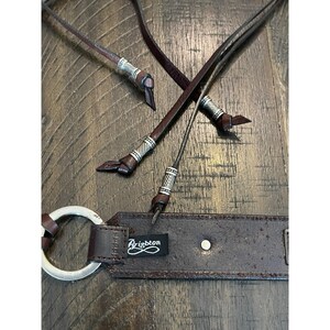 May include: A dark brown leather belt featuring a silver-toned metal ring buckle and thin leather cord accents. The cords are finished with decorative silver-toned metal beads. A black fabric tag with the white text Brighton is attached.