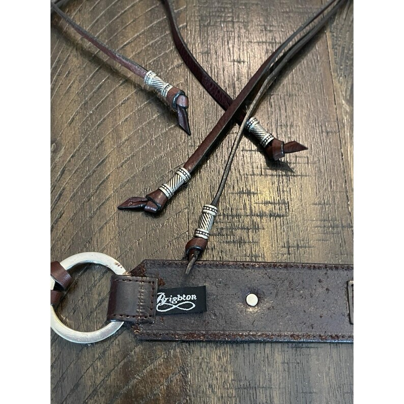 May include: A dark brown leather belt featuring a silver-toned metal ring buckle and thin leather cord accents. The cords are finished with decorative silver-toned metal beads. A black fabric tag with the white text Brighton is attached.