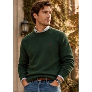 May include: A dark green long-sleeve crewneck sweater with a small red embroidered logo on the chest, worn over a white collared shirt. The sweater features ribbed cuffs and a ribbed hem, paired with blue denim jeans and a brown leather belt.