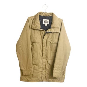 May include: Tan-colored jacket with a dark blue collar and lining. The jacket has four front pockets, a button-up closure, and a small logo on the collar. The jacket is hanging on a metal hanger.