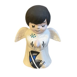 May include: A small white ceramic angel figurine with black hair, closed eyes, and painted details. The angel is wearing a white robe with blue and black accents and has wings.