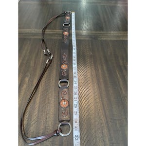 May include: A dark brown leather belt featuring silver-toned metal rings and orange floral embroidery. The belt is positioned next to a white measuring tape, showing a total length of approximately 32 inches from the top to the bottom edge.