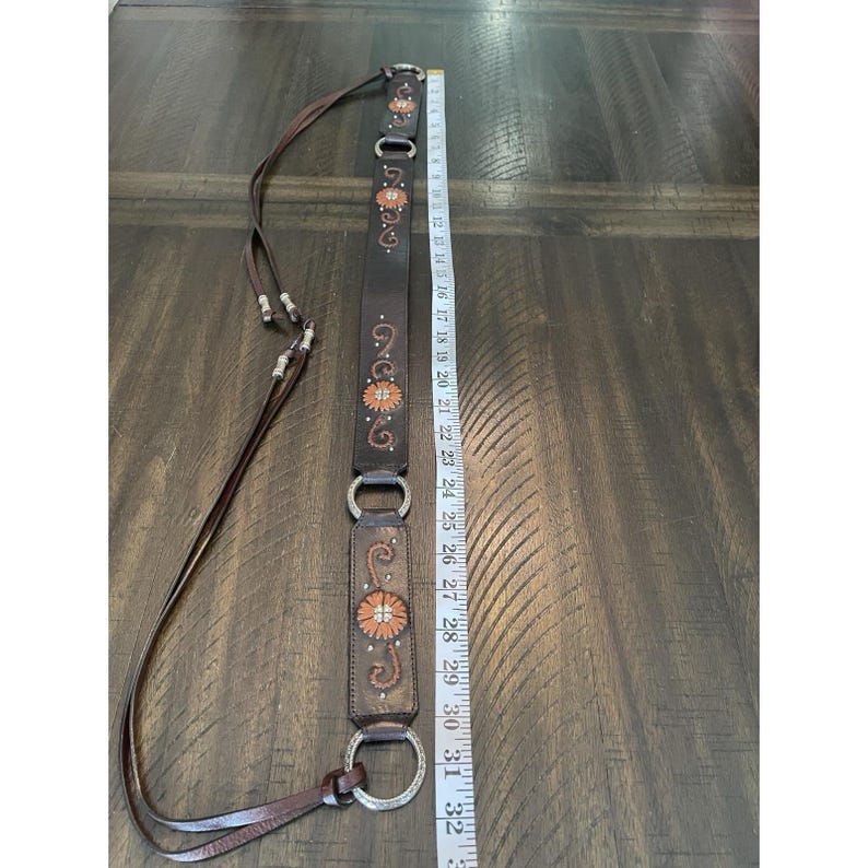 May include: A dark brown leather belt featuring silver-toned metal rings and orange floral embroidery. The belt is positioned next to a white measuring tape, showing a total length of approximately 32 inches from the top to the bottom edge.
