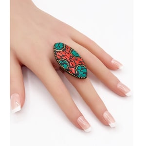 May include: An oval-shaped ring with a mosaic design. The ring features a red and turquoise pattern with a dark metal border. The ring is worn on a finger, showcasing its intricate details and vibrant colors.