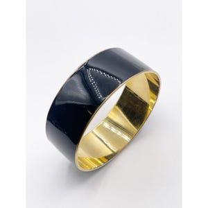 Op de afbeelding: Een brede, glanzend zwarte armband met een goudkleurig interieur. De armband heeft een glad, reflecterend oppervlak en een eenvoudig, elegant ontwerp. Het gouden interieur voegt een vleugje luxe toe aan het accessoire.
