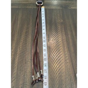 May include: A dark brown leather tassel accessory featuring four thin leather strips attached to a silver-toned metal ring. Each strip ends with a silver-toned metal bead. A white measuring tape shows the length is approximately 19 inches.