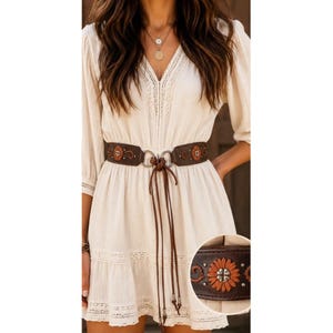 May include: A cream-colored dress with three-quarter sleeves and lace trim details, paired with a wide dark brown leather belt. The belt features a central tie closure with long leather cords and decorative floral embroidery with metal studs.