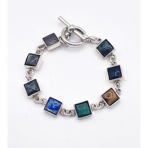 May include: A silver bracelet featuring a series of square-cut gemstones in shades of blue, green, and brown. The bracelet has a toggle clasp and is displayed against a white background.