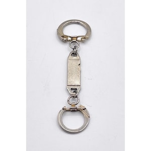 May include: A silver-toned keychain with a rectangular, patterned center piece. The keychain has two key rings, one at the top and one at the bottom, connected by a series of small links. The center piece has a decorative design.