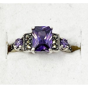 Può includere: Anello d'argento con una grande pietra viola rettangolare al centro. Due pietre viola più piccole affiancano la pietra principale, con dettagli decorativi. L'anello ha uno stile vintage.