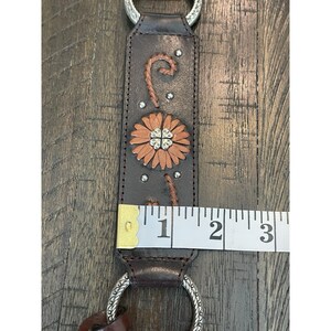 May include: A dark brown leather strap featuring a tan flower applique with a silver-tone metal center, silver studs, and decorative stitching. A white measuring tape shows the strap width is approximately 1.5 inches, with silver rings at both ends.