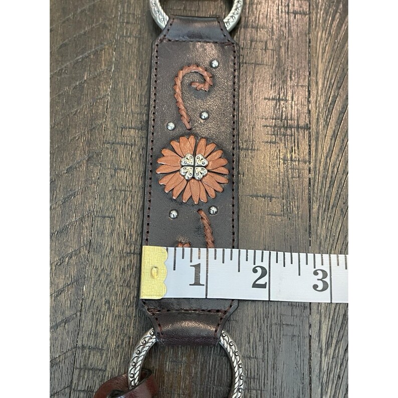 May include: A dark brown leather strap featuring a tan flower applique with a silver-tone metal center, silver studs, and decorative stitching. A white measuring tape shows the strap width is approximately 1.5 inches, with silver rings at both ends.