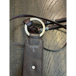 May include: A dark brown leather belt featuring a silver-toned metal ring buckle. The belt strap displays the printed text B30328 and the letter L. It includes thin leather ties accented with small silver-toned metal beads at the ends of the strap.