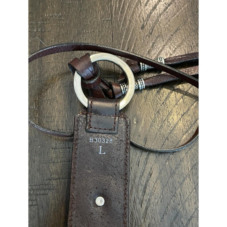 May include: A dark brown leather belt featuring a silver-toned metal ring buckle. The belt strap displays the printed text B30328 and the letter L. It includes thin leather ties accented with small silver-toned metal beads at the ends of the strap.
