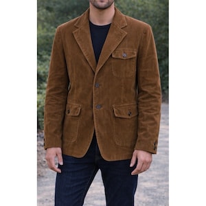 Puede incluir: Blazer de pana marrón con cuatro bolsillos delanteros y cierre de dos botones. El blazer tiene solapas con muescas y se lleva sobre una camiseta negra. El modelo también lleva vaqueros azul oscuro.