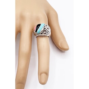 May include: A silver ring with a colorful stone inlay. The stone features black, pink, and turquoise sections. The ring has an ornate, openwork design around the stone. The ring is shown on a finger.
