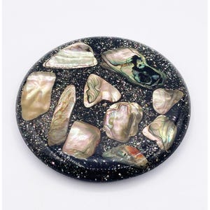 Puede incluir: Una pieza decorativa redonda y negra con incrustaciones de conchas de abulón iridiscentes. Las conchas varían en tamaño y forma, mostrando tonos de blanco nacarado, rosa y verde. La base negra está salpicada de purpurina. Adecuado para la decoración del hogar.