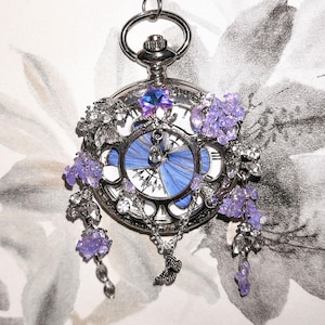 Peut inclure: Un pendentif de montre de poche argenté avec un motif d'aile de papillon bleu sur le cadran. La montre est ornée de fleurs violettes et de cristaux transparents. Un petit papillon argenté est suspendu.