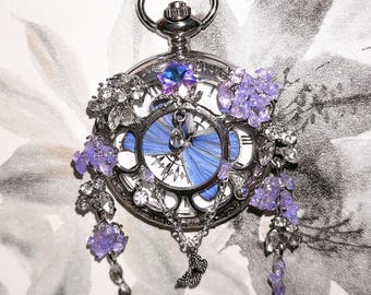 May include: A silver pocket watch pendant with a blue butterfly wing design on the face. The watch is adorned with purple floral accents and clear crystals. A small silver butterfly charm hangs from the watch.