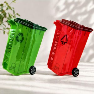 May include: Two miniature, decorative trash cans. One is bright green with a recycling symbol, the other is red with a recycling symbol. Both have black wheels and dark green or brown lids. The cans are leaning slightly.