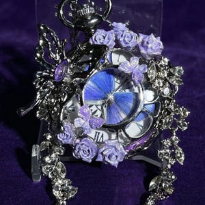 May include: A silver-toned pocket watch with a butterfly and floral design. The watch face features a blue butterfly design. Lavender roses and crystals adorn the watch's frame. The watch is displayed on a purple surface.
