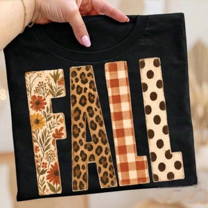May include: A black t-shirt featuring the word FALL in large block letters. Each letter has a distinct pattern: the F has a floral print, the A has a leopard print, the L has a brown and white gingham check, and the second L has brown polka dots.