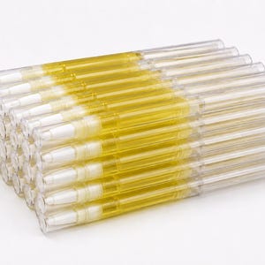 Könnte beinhalten: Ein Stapel aus durchsichtigen Kunststoff-Drehstiften, die mit einer transparenten gelben Flüssigkeit gefüllt sind. Jeder Stift hat an einem Ende eine weiße Pinselspitze und am anderen eine durchsichtige Kappe, angeordnet in einem rechteckigen Raster.