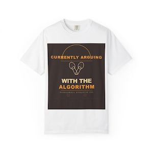 May include: White t-shirt with a dark brown square graphic. The graphic features the text "CURRENTLY ARGUING WITH THE ALGORITHM" in white and orange, along with pickleball paddles. The shirt is from Pickleball Athletic Co.