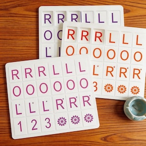Könnte beinhalten: Drei weiße Buchstabenkarten mit lila, orangefarbenen und roten Buchstaben. Die Karten haben Buchstaben, Zahlen und Blumendesigns. Die Karten liegen auf einer Holzoberfläche. Eine kleine blaue Schale befindet sich unten rechts.