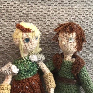 May include: Two handmade crochet dolls featuring yarn hair and plastic eyes. The left doll has blonde hair, a green top, brown skirt, and tan arm cuffs. The right doll has brown hair, a green tunic with a brown collar, and dark green pants.