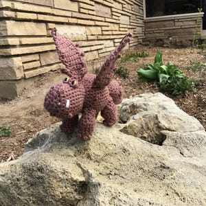 May include: A crocheted dragon toy in a dusty rose color, with large wings, small yellow eyes, and a white tooth. The dragon is standing on a large rock outdoors, with a brick building in the background.